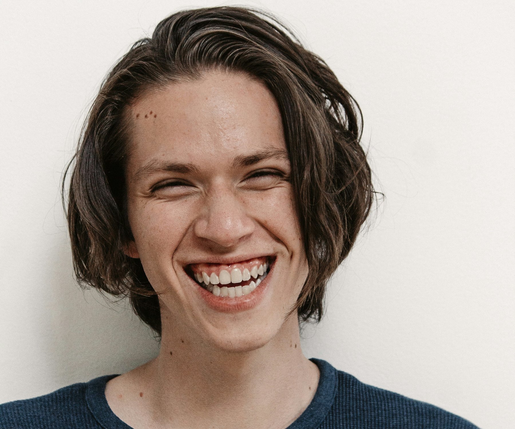 Young man with shoulder-length hair laughing, in a navy sweater against a plain light wall